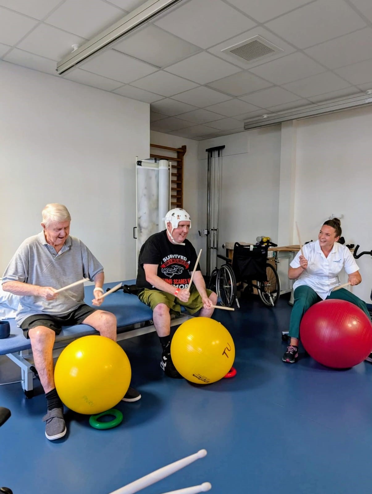 Music physiotherapy 2 men and female physiotherapist banging drumsticks on large yellow and red balls