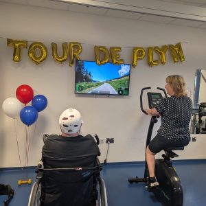 Man in wheelchair with white helmet on hand bike with woman on exercise bike. Balloons spell out Tour de Plym on wall.