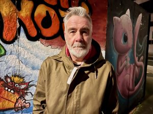 Man in khaki jacket stood by a wall of grafitti