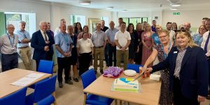 Two women cutting cake in front of group of people stood in patient lounge 