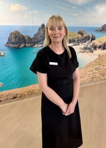 Woman with blonde hair wearing black dress, stood in front of a mural of a sandy cove