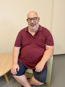 Man sat in chair with glasses and grey beard. Wearing burgundy t-shirt and blue shorts.