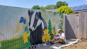 Man sat on floor spraying fence artwork featuring a badger and flowers 