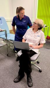 Nurse in blue uniform stood by podiatrist in white clinical uniform sat down with a laptop.