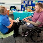 Woman in blue t-shirt sat down taking blood pressure of a male wheelchair user wearing burgundy t-shirt