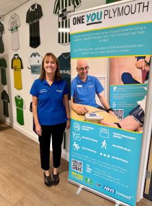 Woman in blue t-shirt and black trousers stood by One You Plymouth health banner